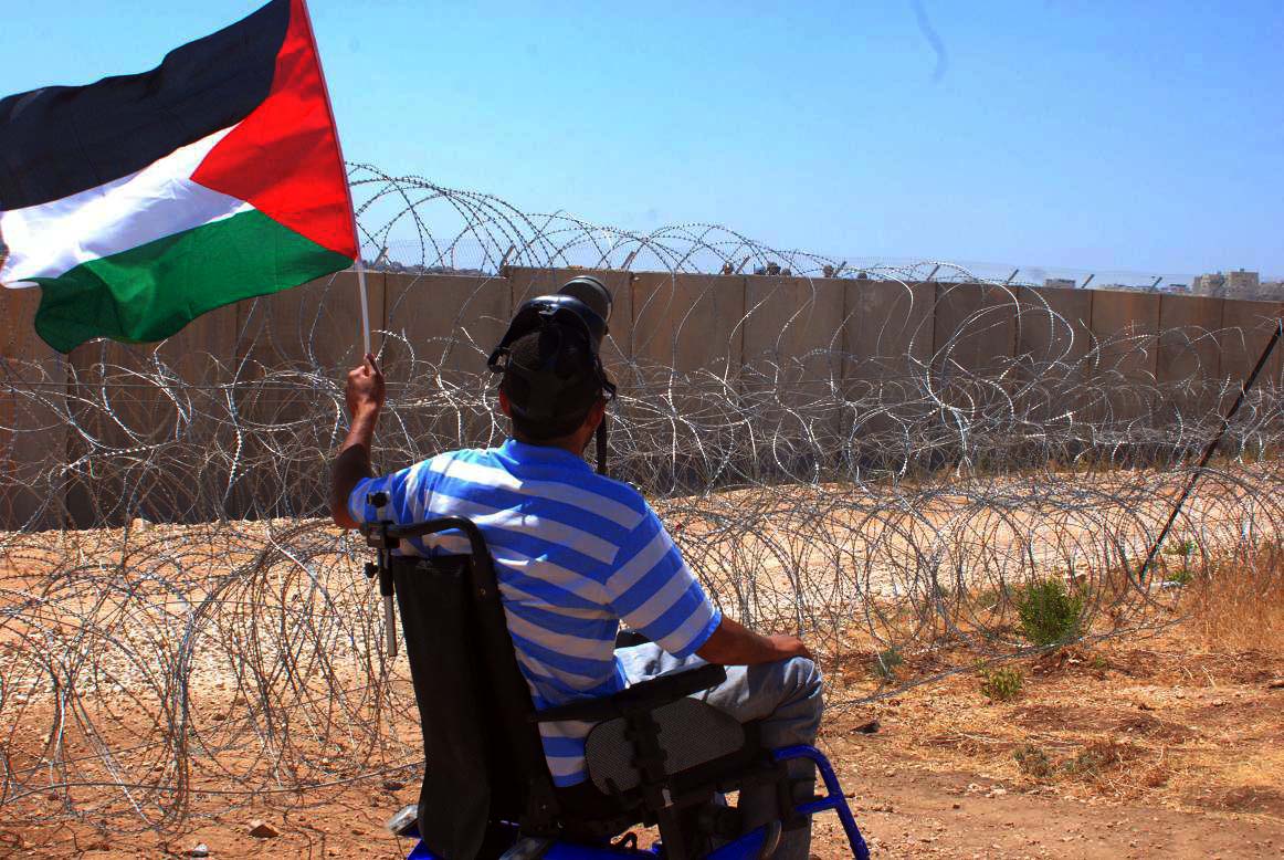 Мужчина-колясочник держит в руках флаг Палестины около разделительной стены, возведенной Израилем на палестинских территориях 
